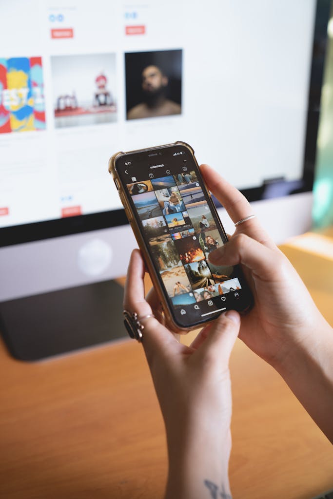 Close-up of a person browsing a smartphone with an Instagram feed, with a computer monitor in the background.